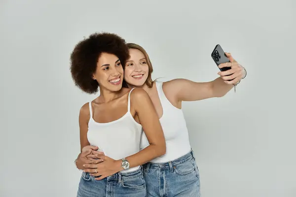 Two women, one with curly hair and one with straight hair, wearing white tank tops and jeans, pose for a selfie. — Stock Photo