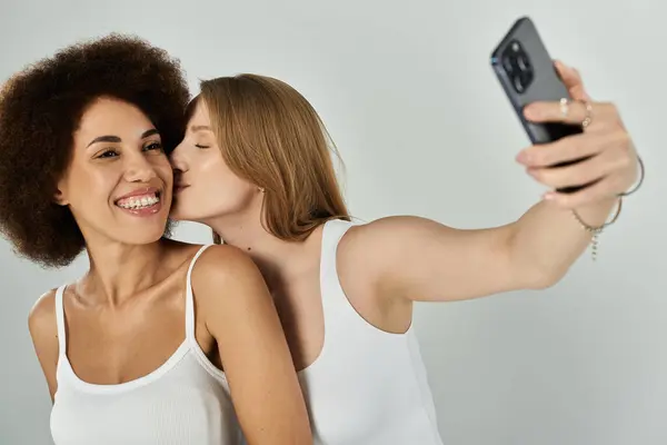 Un couple de lesbiennes noires et blanches en débardeurs et en jeans prend un selfie, embrassant la joue — Photo de stock