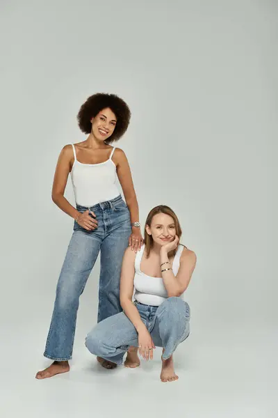 Due donne, una nera e una bianca, che indossano jeans e canottiere bianche posano insieme su uno sfondo grigio. — Foto stock