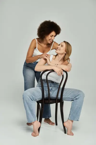 A lesbian couple in white tank tops and jeans share a loving gaze while sitting on a chair in a studio setting. — Stock Photo
