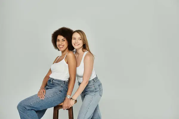 A lesbian couple, one Black and one white, are smiling at the camera while wearing white tank tops and jeans against a gray backdrop. — Stock Photo