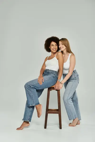 Dos mujeres, una negra y otra blanca, en jeans y camisetas blancas se sonríen. Mujer en el taburete tiene pies en el suelo — Stock Photo