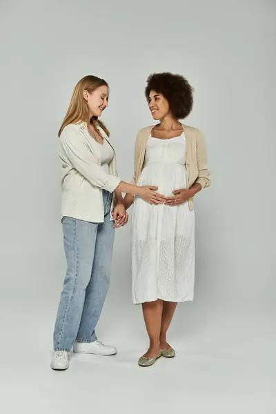 Una mujer afroamericana embarazada con un vestido blanco sonríe a su pareja, que toca suavemente su vientre. - foto de stock