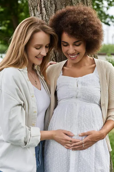 Two women, one pregnant, stand close together under a tree, hands resting on the pregnant womans belly. — Stock Photo