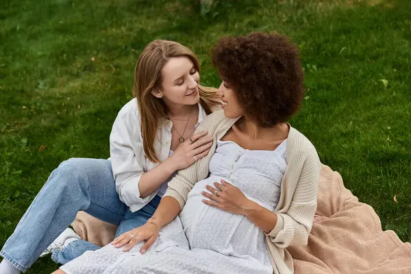 A pregnant woman rests in the grass, her partner cradles her arm as they gaze at each other lovingly. — Stock Photo