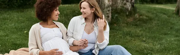 Two women, one pregnant, sit on a blanket in a grassy park, smiling and holding hands. — Stock Photo