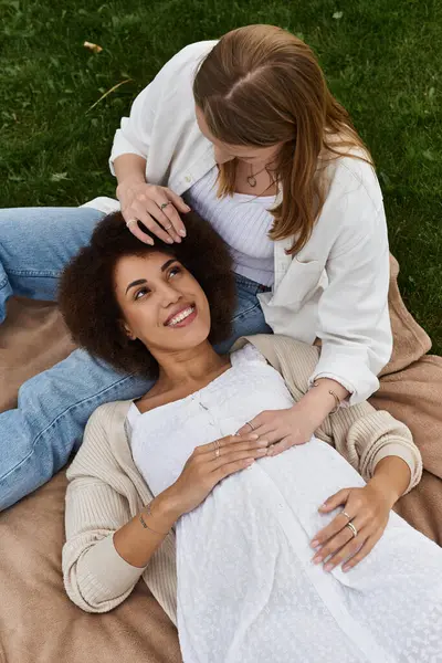 Eine schwangere Frau liegt auf dem Rücken in einem Park, ihr Partner legt eine Hand auf ihren Bauch. Beide Frauen lächeln glücklich. — Stockfoto