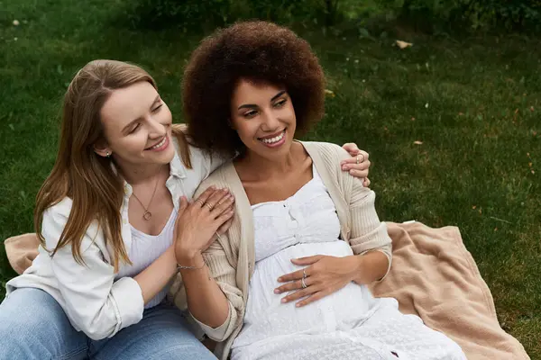 Una donna incinta, cullandosi la pancia, sorride amorevolmente mentre la sua compagna poggia una mano sulla spalla. — Foto stock