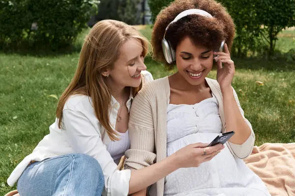 Two women, one pregnant, sit on a blanket in a grassy park, smiling and listening to music on headphones. — Stock Photo