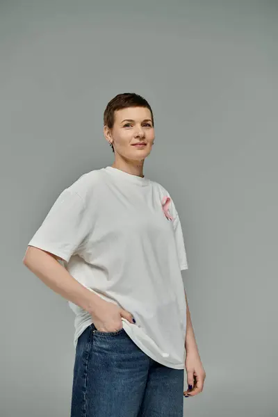 A woman with short hair, wearing a white t-shirt with a pink ribbon, stands confidently against a grey backdrop. — Stock Photo
