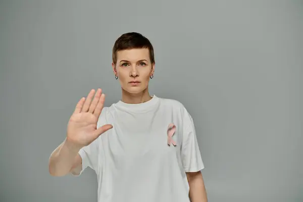 Женщина с короткими волосами и розовой лентой на рубашке протягивает руку в знак 