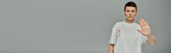 Una donna con i capelli corti e un nastro rosa sulla camicia alza la mano, palmo rivolto verso la macchina fotografica, su uno sfondo grigio. — Foto stock
