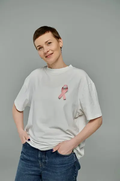 A woman with short hair and a pink ribbon on her shirt stands against a grey background. — Stock Photo