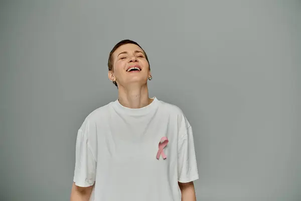 Una mujer con el pelo corto, con ropa casual y una cinta rosa, sonríe brillantemente sobre un fondo gris. - foto de stock