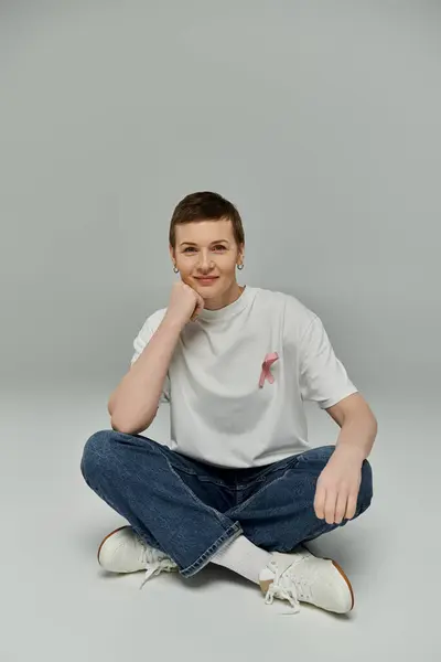 A woman with short hair wearing casual attire sits on the floor with her legs crossed, displaying a pink ribbon on her shirt. — Stock Photo