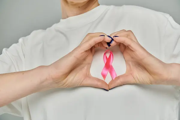Short-haired woman in white shirt forms heart with hands, emphasizing pink ribbon for breast cancer awareness — Stock Photo