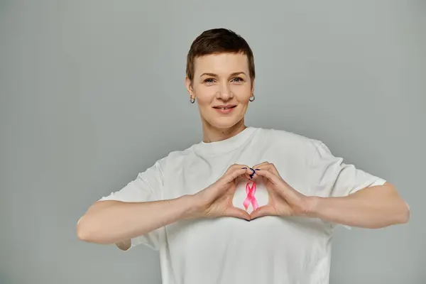 Woman with short hair, white shirt and pink ribbon forms a heart with hands, showing support for breast cancer awareness — Stock Photo