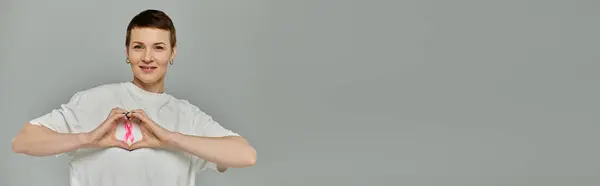 Woman in short hair and white T-shirt forms heart with hands, holding pink ribbon as symbol for breast cancer awareness — Stock Photo