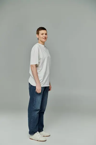 Mujer de pelo corto en jeans, camiseta blanca y zapatillas de deporte sonríe con confianza contra un telón de fondo gris en atuendo casual - foto de stock