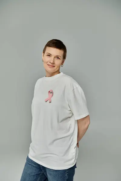 A woman with short hair stands against a gray backdrop, wearing a white t-shirt with a pink ribbon pinned to it. — Stock Photo