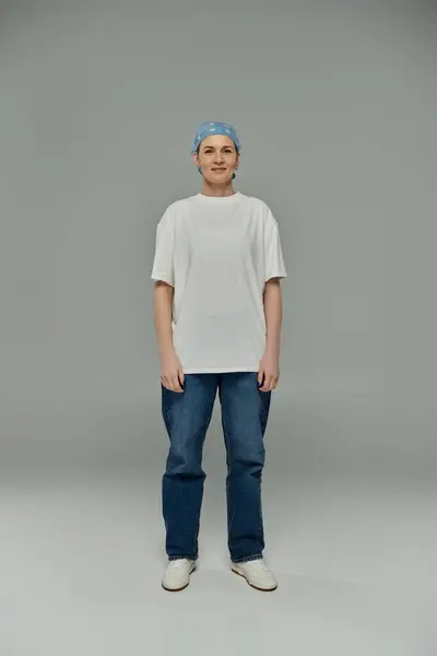 A woman with short hair, wearing a white t-shirt, blue jeans, stands against a gray background. — Stock Photo