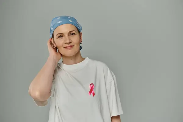 A woman with short hair wears a pink ribbon on her white shirt, standing against a gray background. — Stock Photo