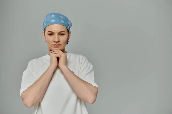 Mulher de cabelos curtos em t-shirt branca, bandana azul, contra fundo cinza, mãos juntas, fita rosa na camisa — Fotografia de Stock