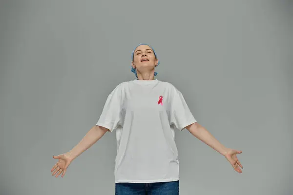 A woman in casual attire with a pink ribbon stands against a grey background, symbolizing breast cancer awareness. — Stock Photo