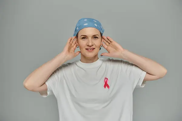 A woman with short hair and a pink ribbon on her shirt smiles, cupping her hands around her ears against a grey backdrop. — Stock Photo