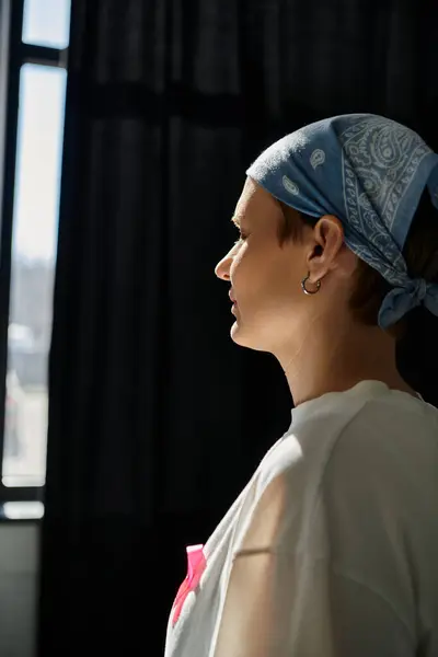 A woman with short hair and a pink ribbon stands in front of a window, looking out with a thoughtful expression. — Stock Photo