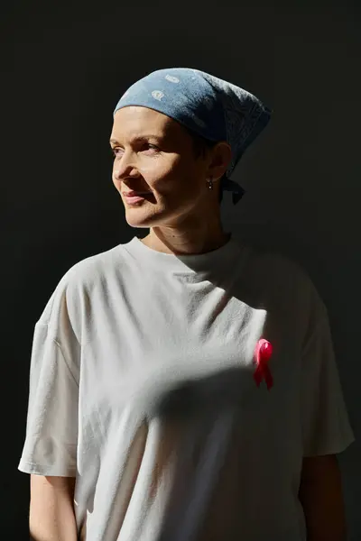 Woman in white t-shirt and blue bandana stands with pink ribbon for breast cancer awareness against grey background — Stock Photo