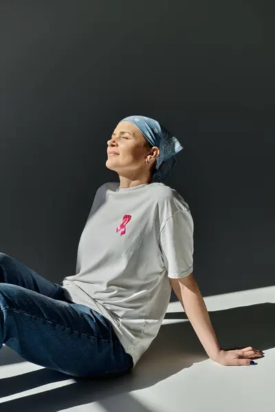 A woman wearing a blue bandana and a pink ribbon on her shirt sits on a grey background, looking upwards. — Stock Photo