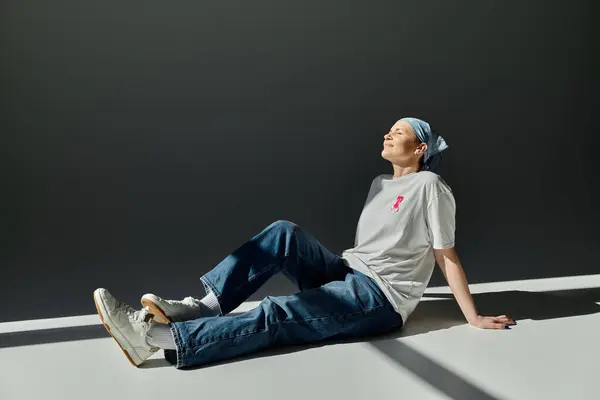Woman sits on white floor in casual attire, pink ribbon, blue head scarf, with sunlight streaming through window — Stock Photo