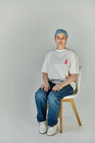 Una donna siede su una sedia bianca, indossando jeans e una t-shirt bianca con un nastro rosa, il che significa consapevolezza del cancro al seno.. — Foto stock