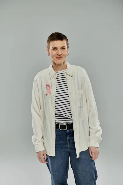 A woman with short hair, wearing casual clothing and a pink ribbon, stands against a grey background. — Stock Photo