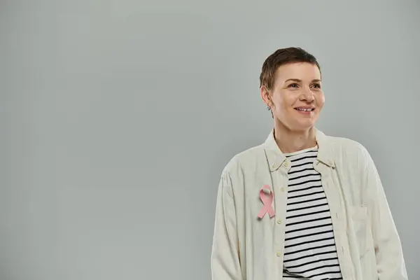 A woman with short hair and a pink ribbon pinned to her shirt smiles and looks to the side. — Stock Photo