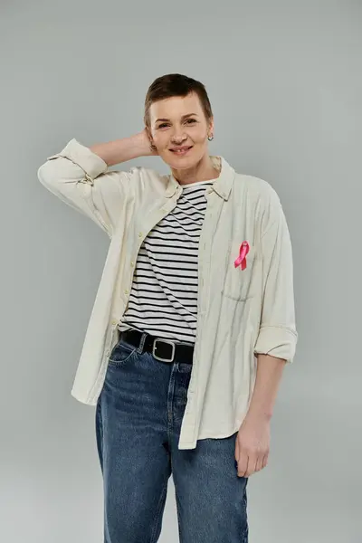 Short-haired woman in white shirt over striped shirt, blue jeans, and black belt, wears pink ribbon on shirt — Stock Photo