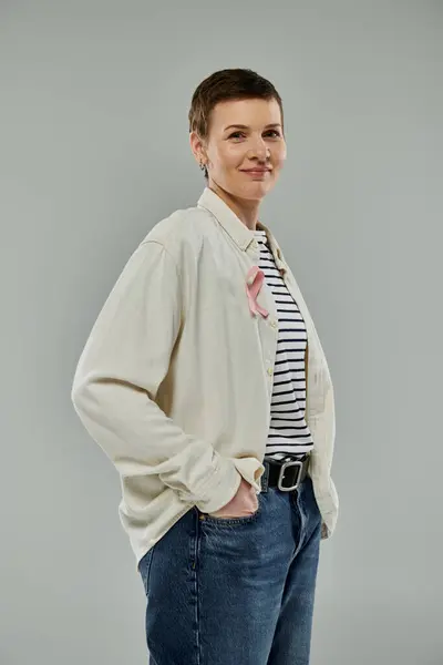 Woman with short hair wears white shirt and jeans in front of gray background. Pink ribbon symbolizes breast cancer awareness — Stock Photo