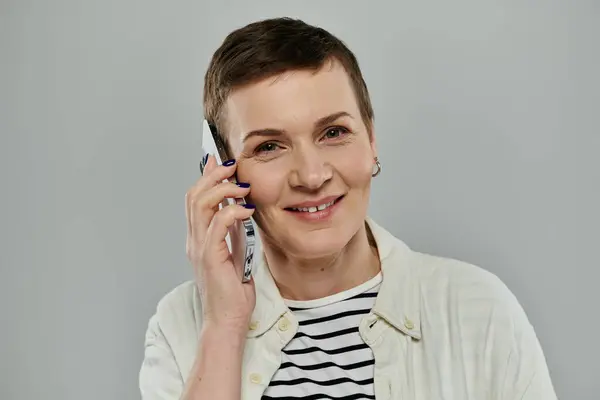 A woman with short brown hair smiles warmly while talking on a smartphone against a gray background. — Stock Photo