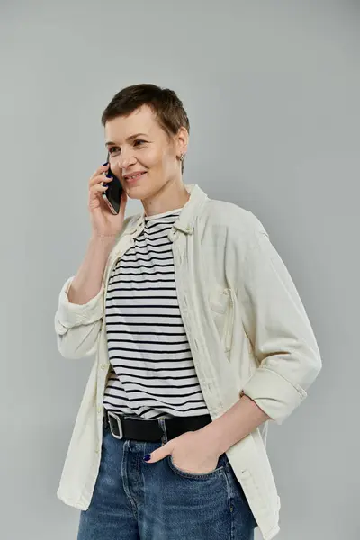 A woman with short hair, wearing a white shirt over a striped tee and jeans, smiles as she talks on her phone. — Stock Photo