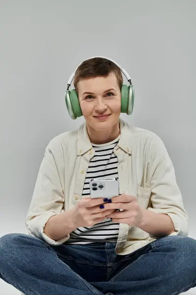 Una mujer con el pelo corto se sienta con las piernas cruzadas, usando auriculares y un teléfono, sobre un fondo gris. — Stock Photo