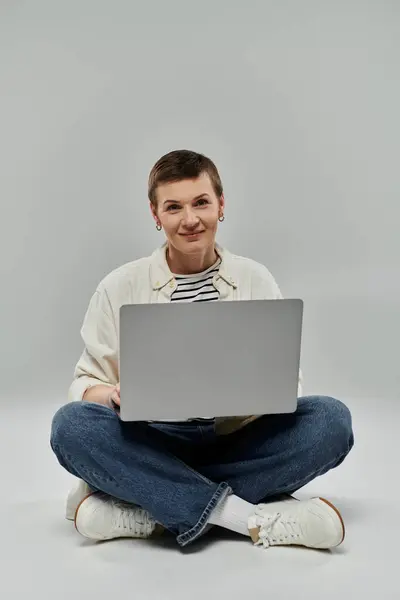 A woman with short hair sits on a grey background, using a laptop and smiling. — Stock Photo