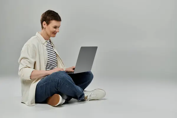 A woman with short hair sits cross-legged on a grey background, using a laptop. — Stock Photo