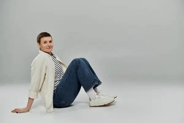 A woman with short hair sits on a grey background, wearing a white shirt and jeans. — Stock Photo