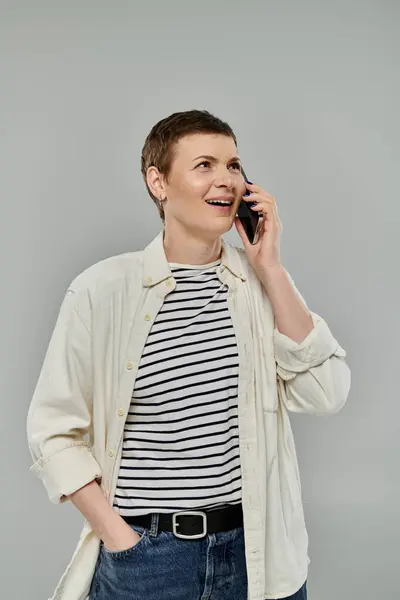 Una donna con i capelli corti, con una camicia bianca sopra una camicia a righe, sorride mentre parla al cellulare. — Foto stock