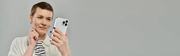 A woman with short hair, wearing casual attire, smiles as she holds a smartphone against a gray backdrop. — Stock Photo