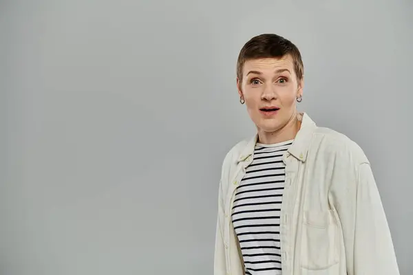 A woman with short hair is surprised and looks to the left of the frame. She is wearing casual attire and standing against a grey backdrop. — Stock Photo
