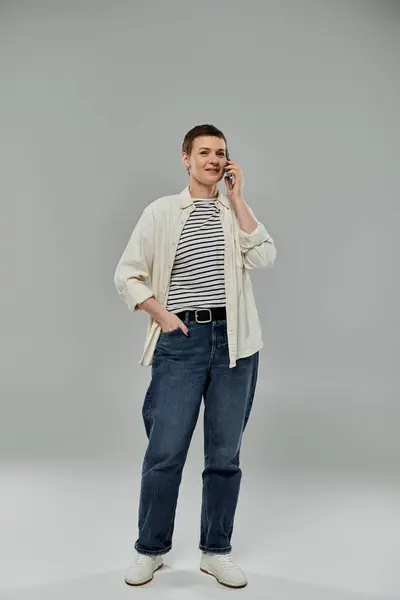 Eine Frau mit kurzen Haaren, lässig in Jeans und Knopfleiste gekleidet, lächelt, als sie vor grauem Hintergrund mit ihrem Handy telefoniert.. — Stockfoto