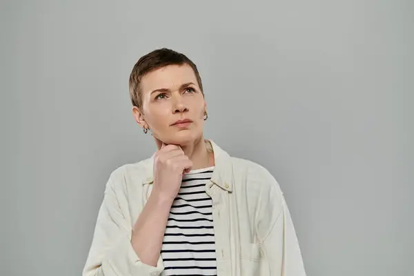 A woman with short hair, wearing a white shirt, ponders thoughtfully against a gray background. — Stock Photo