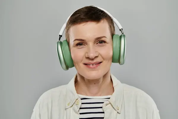 A woman with short hair, wearing casual attire, listens to music on headphones while smiling. She is on a gray background. — Stock Photo